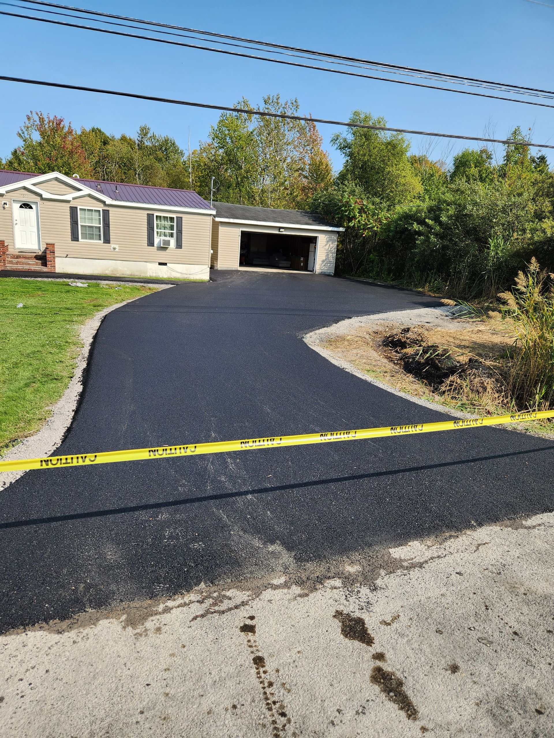 Fresh black asphalt driveway leading to a tan house with a garage, bordered by trees and shrubs