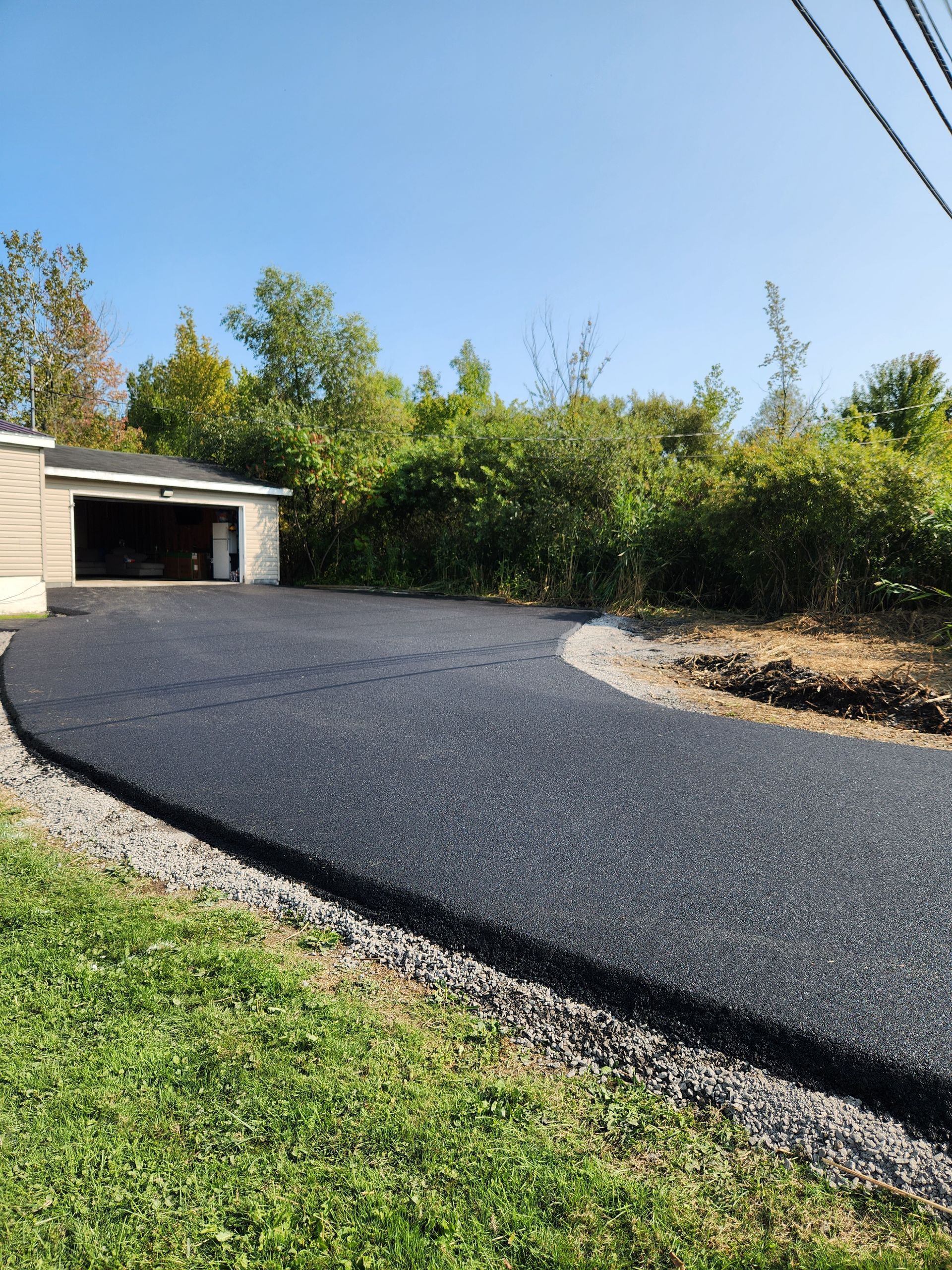 Fresh black asphalt driveway leading to a garage, bordered by gravel, grass, and trees under a blue sky