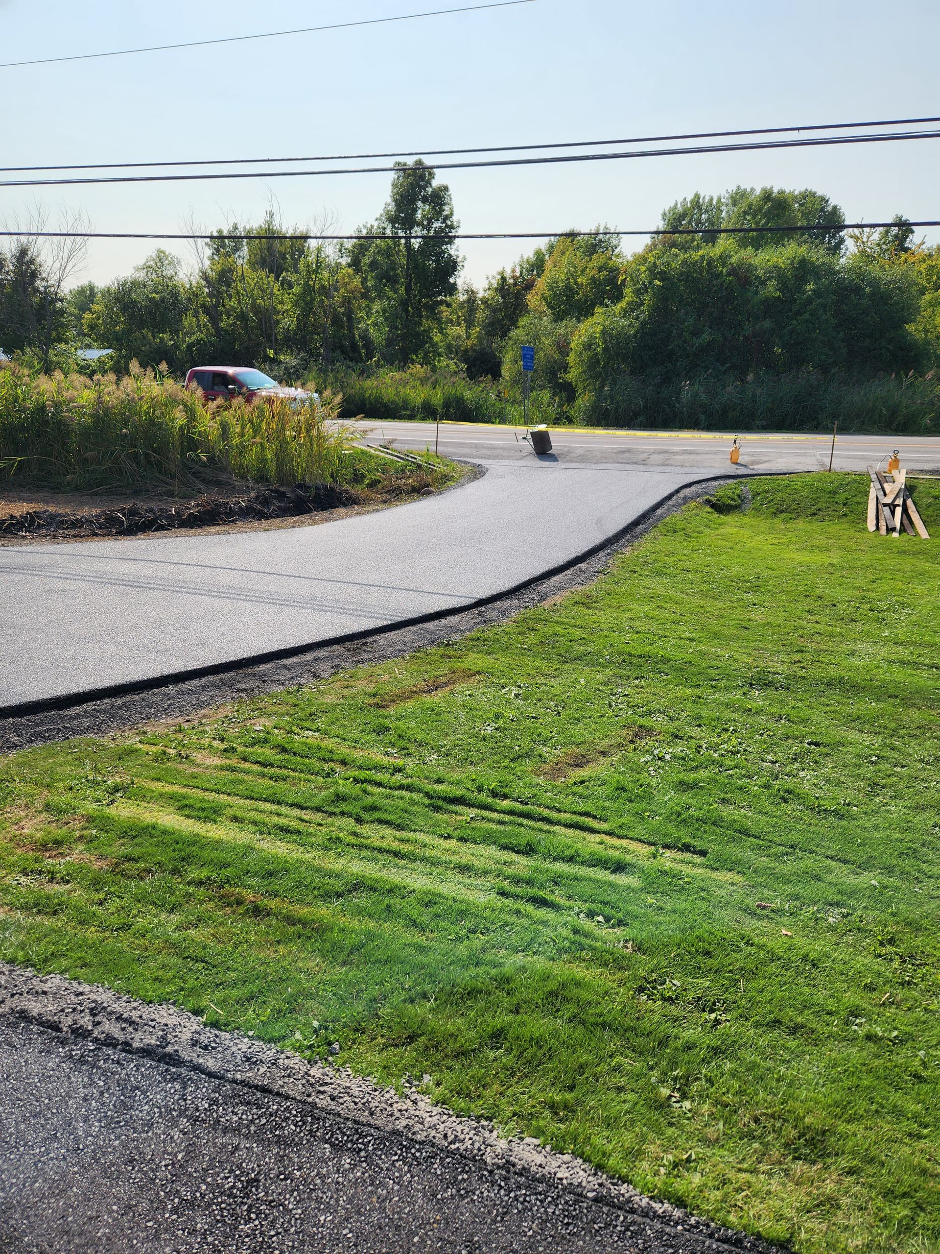 Gravel path through a green lawn and shrubs, with a small black cat sitting on the path near a parked car.