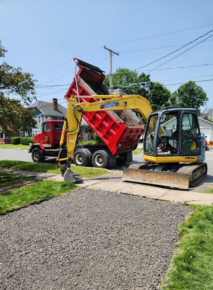 Backhoe loading a red dump truck at a sidewalk work site on a sunny street