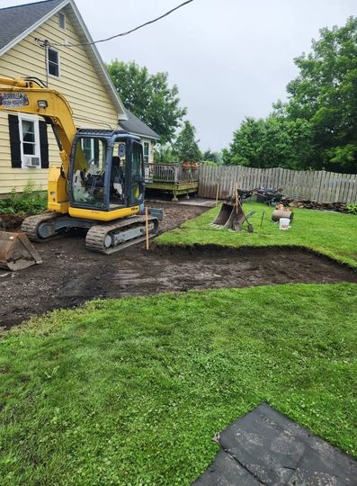 Backyard excavation beside a yellow house, with a mini excavator and fresh soil beds.