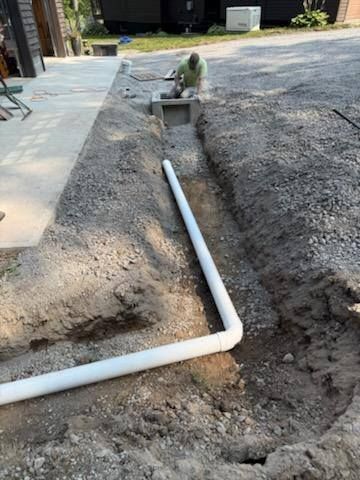 White pipe trench in a gravel yard beside a walkway, with a worker fitting the pipe.