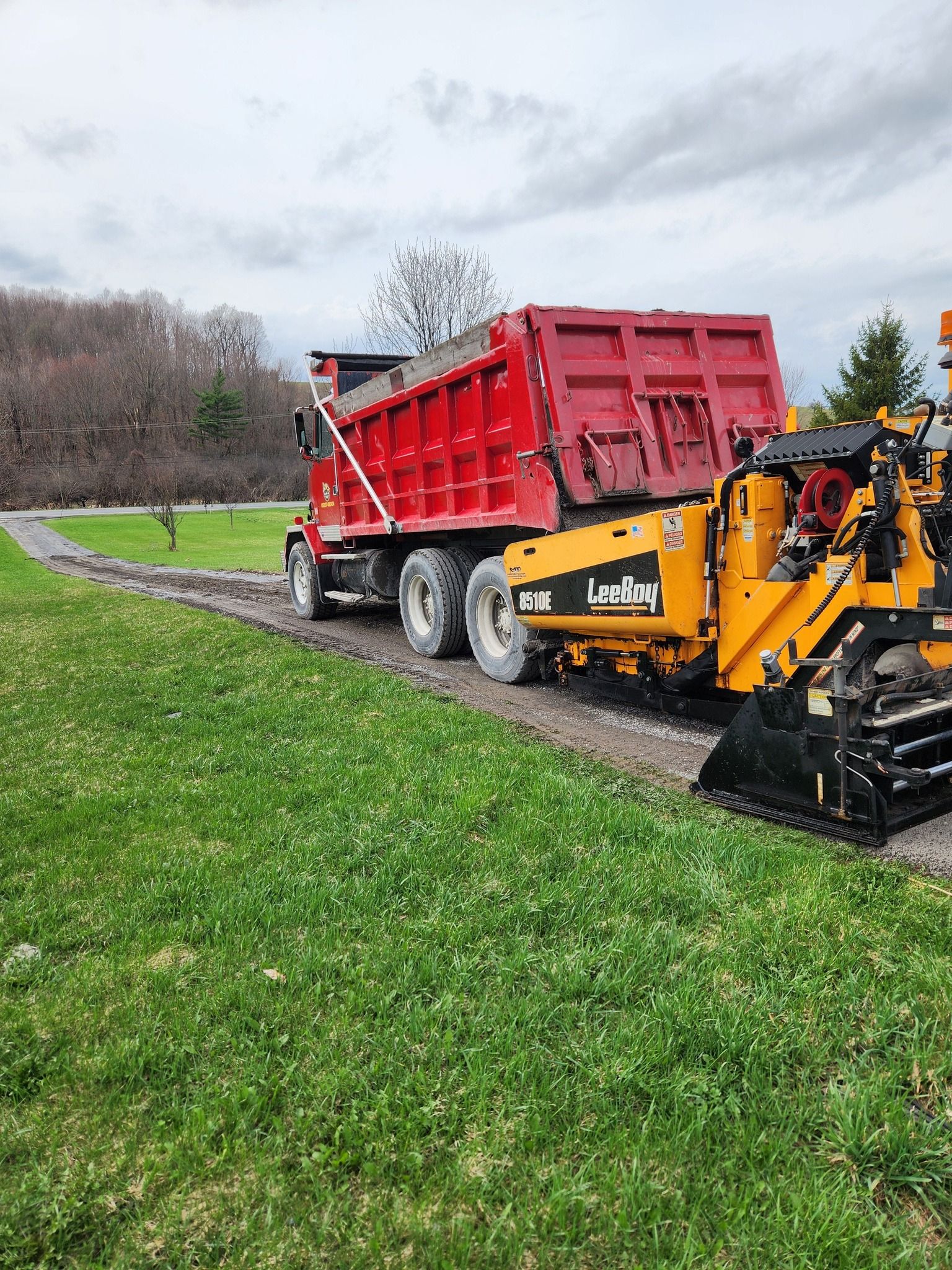 Red dump truck and yellow asphalt machine working on a wet roadside path in a park.