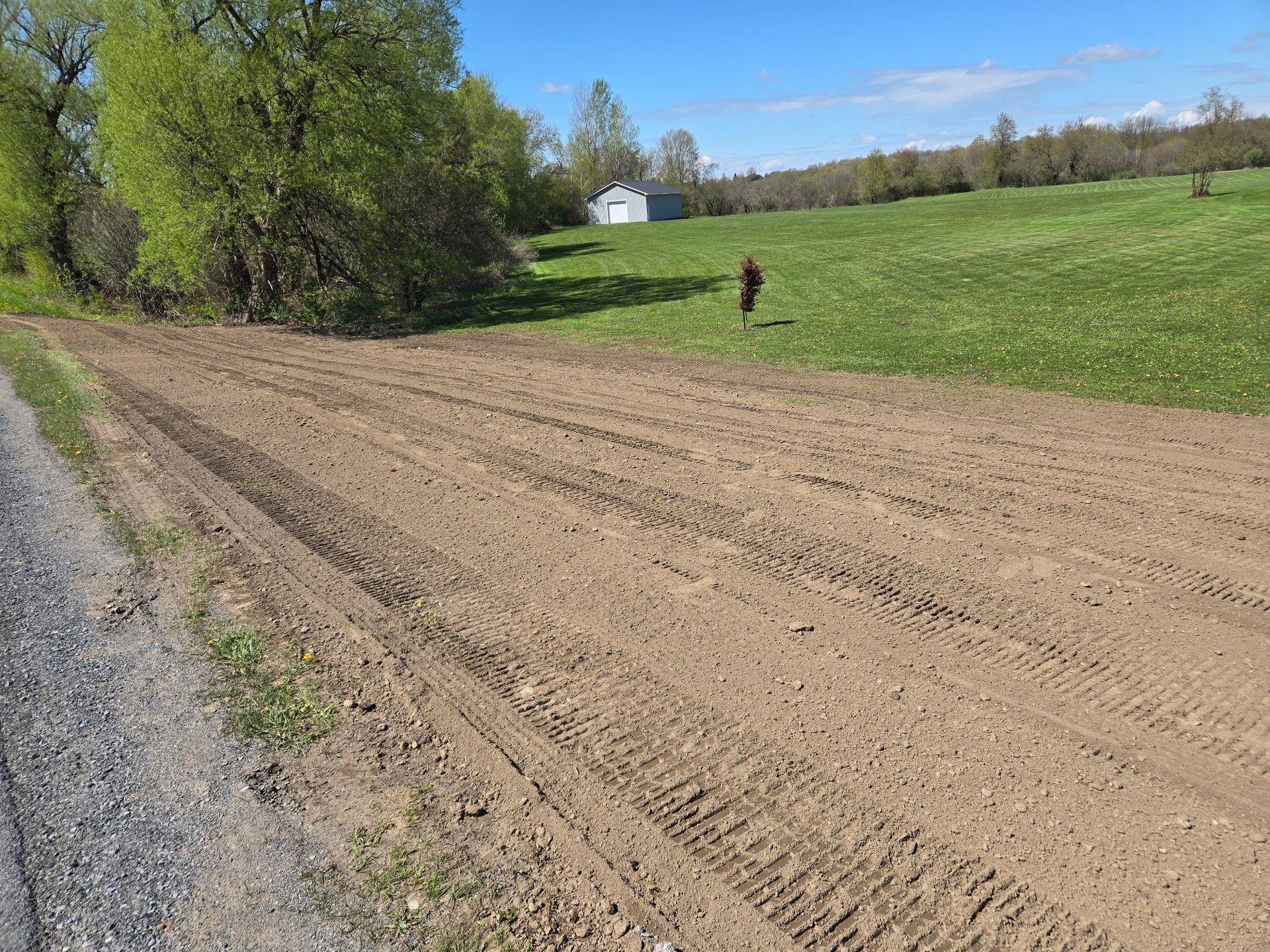 Plowed dirt field beside a gravel road, with green hillside, trees, and a person in the distance
