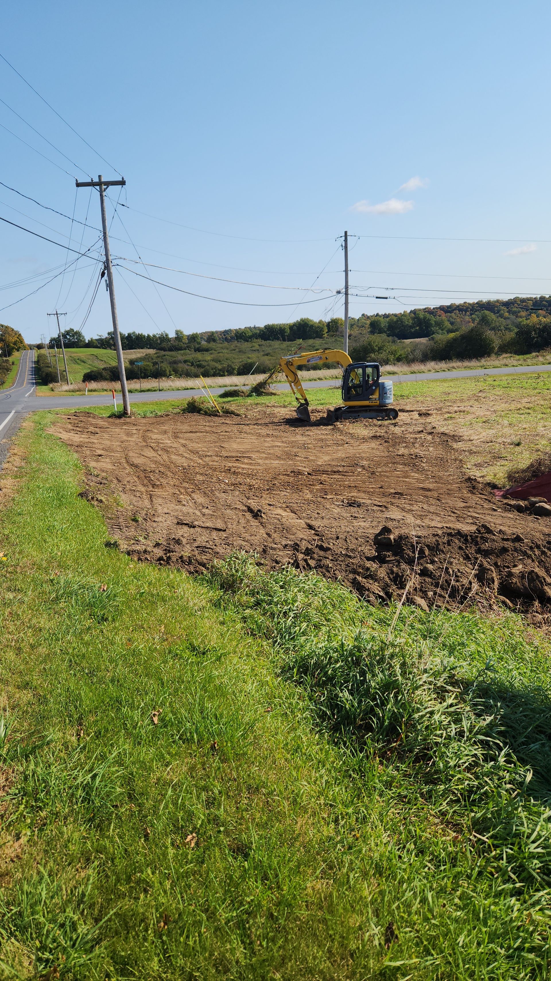 A small tractor tills a field beside a grassy roadside under a clear blue sky.