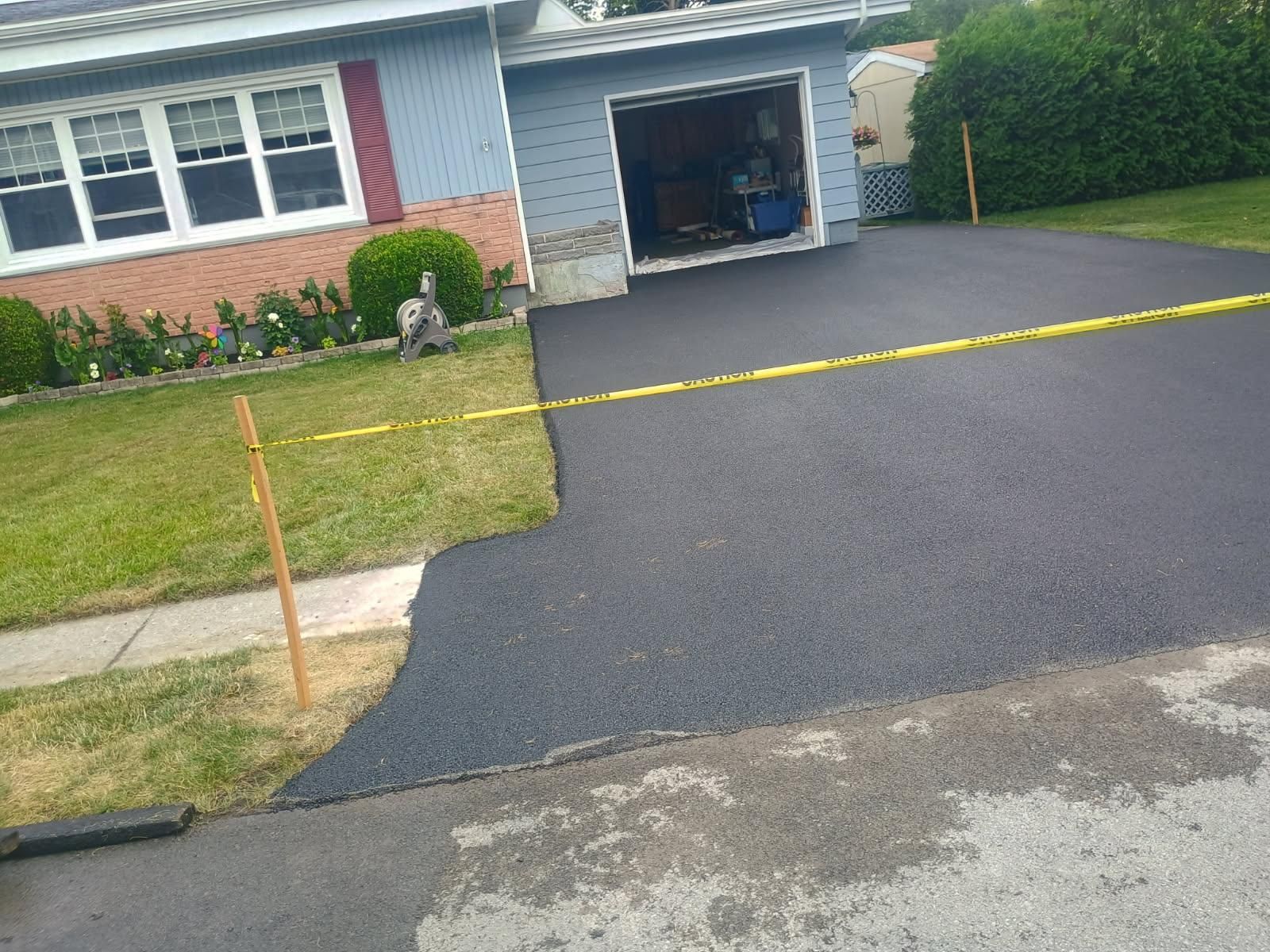 Freshly paved driveway beside a brick house, with a yellow caution line across the entrance.
