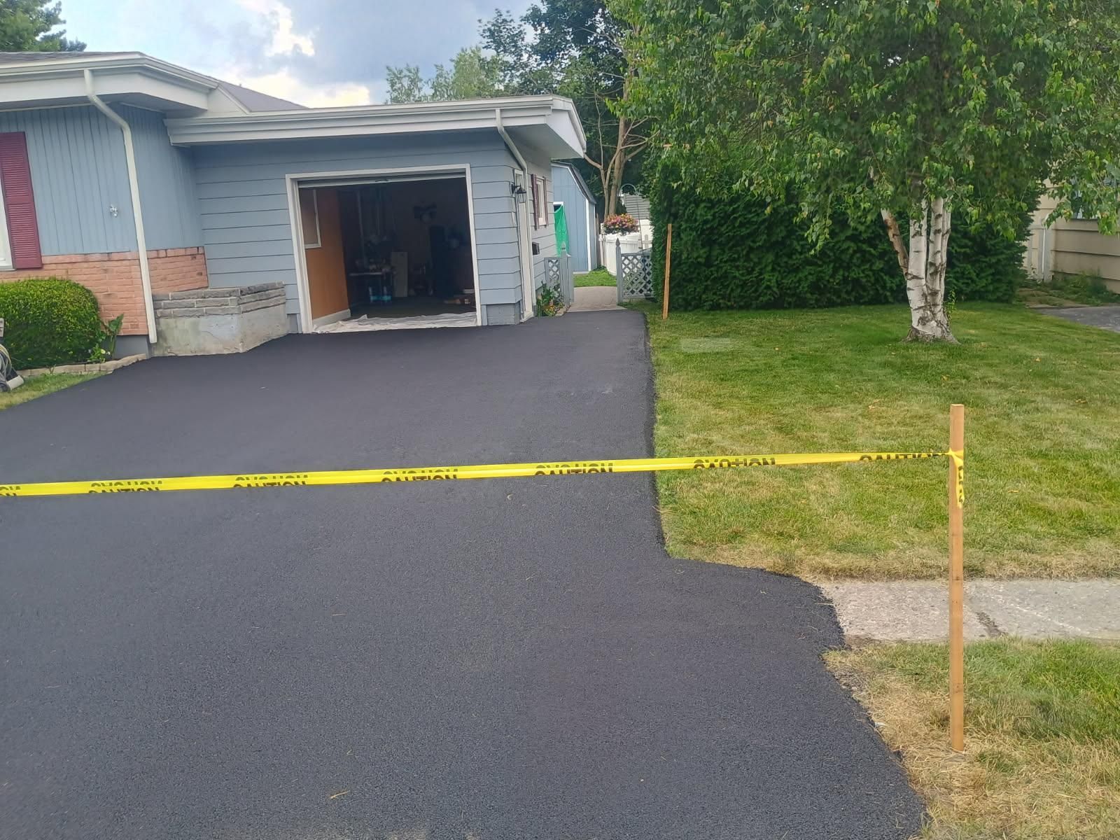 Driveway with fresh black asphalt leading to an open garage, bordered by yellow caution tape and trees.