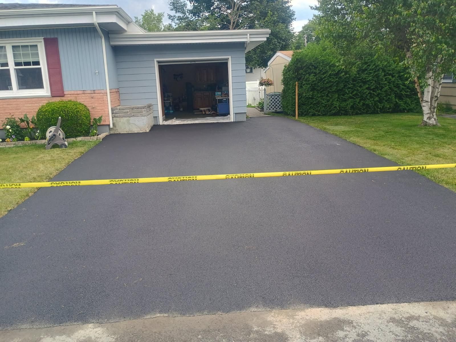 Freshly paved black driveway leading to a blue house with an open garage, marked by yellow caution tape