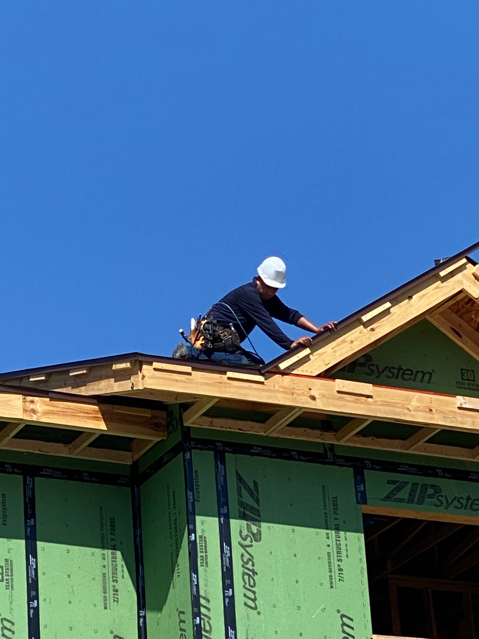Construction worker on roof, wearing hard hat, installing shingles, against a blue sky.
