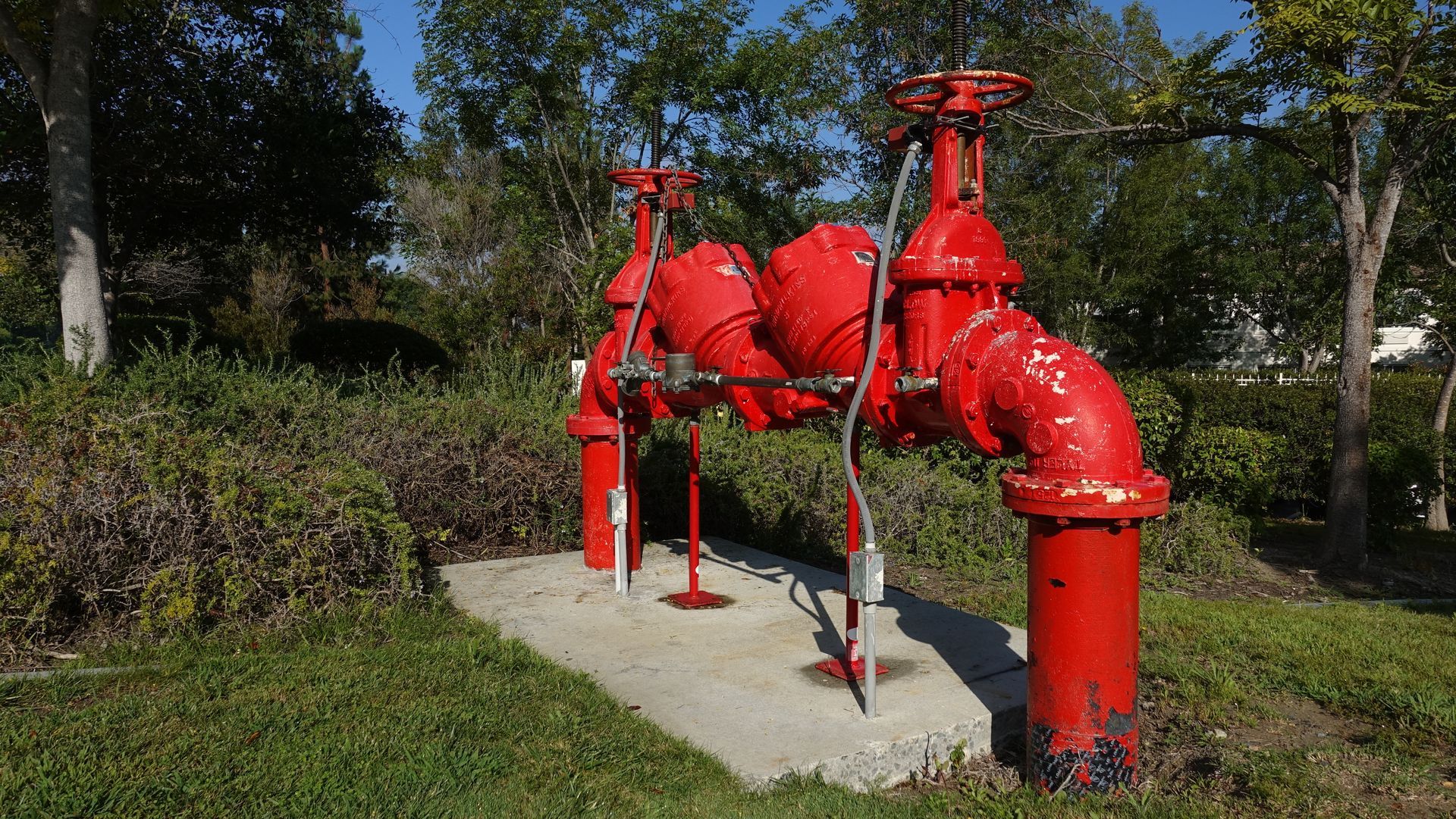 A red, dual-valve pipe assembly mounted on a concrete pad outdoors, surrounded by green landscaping and trees.