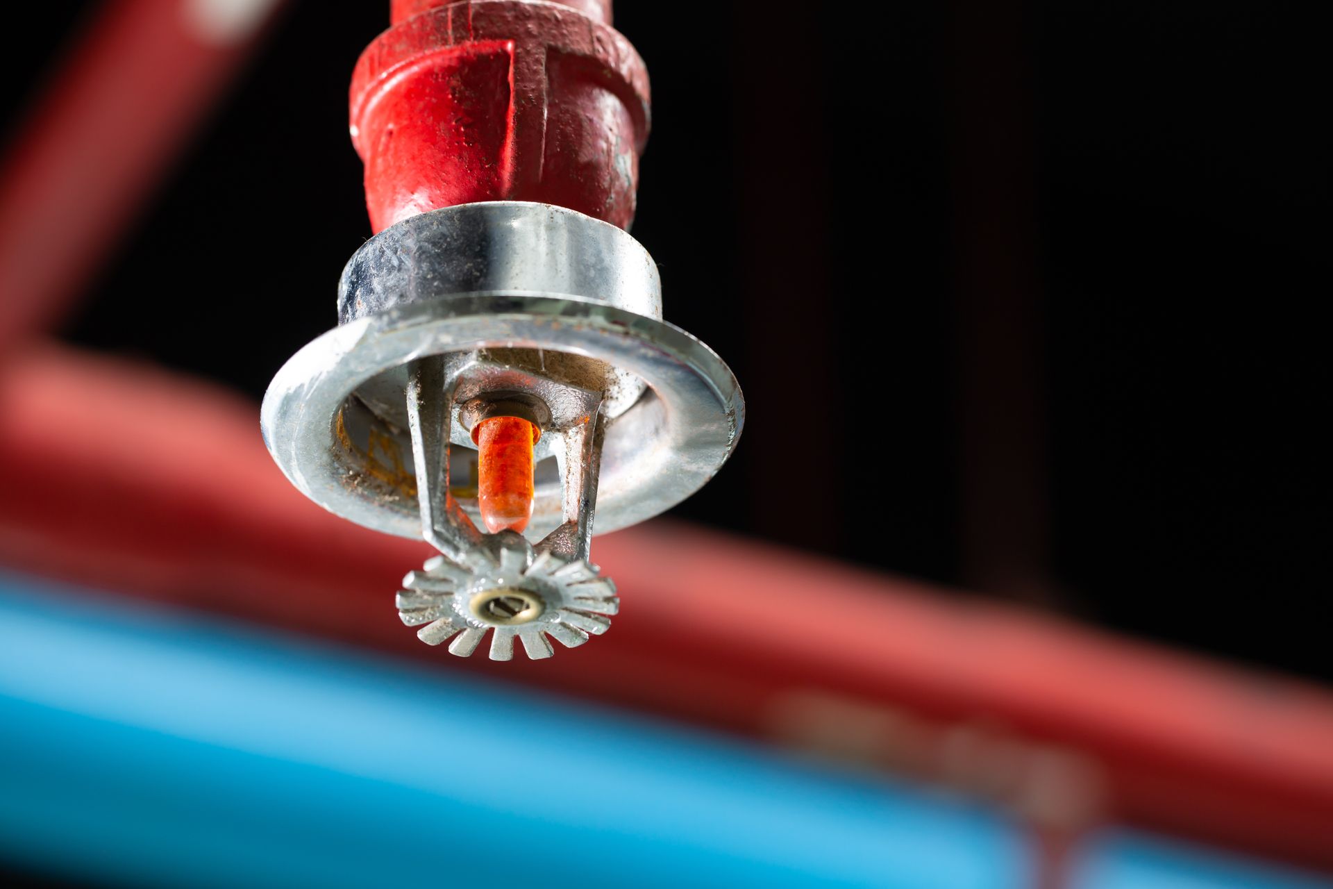 Close-up of a red fire sprinkler head with a shiny metal guard and a red pipe in a dark setting.