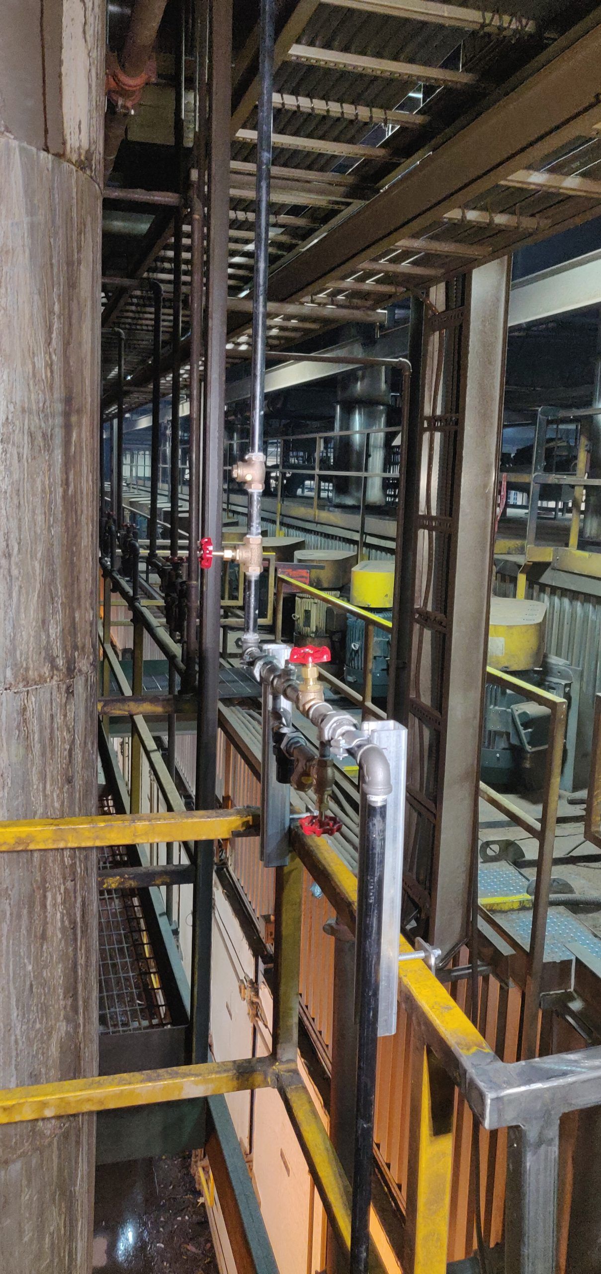 An industrial interior featuring pipes, valves, and a yellow-railed walkway. Metal framework and large tanks.