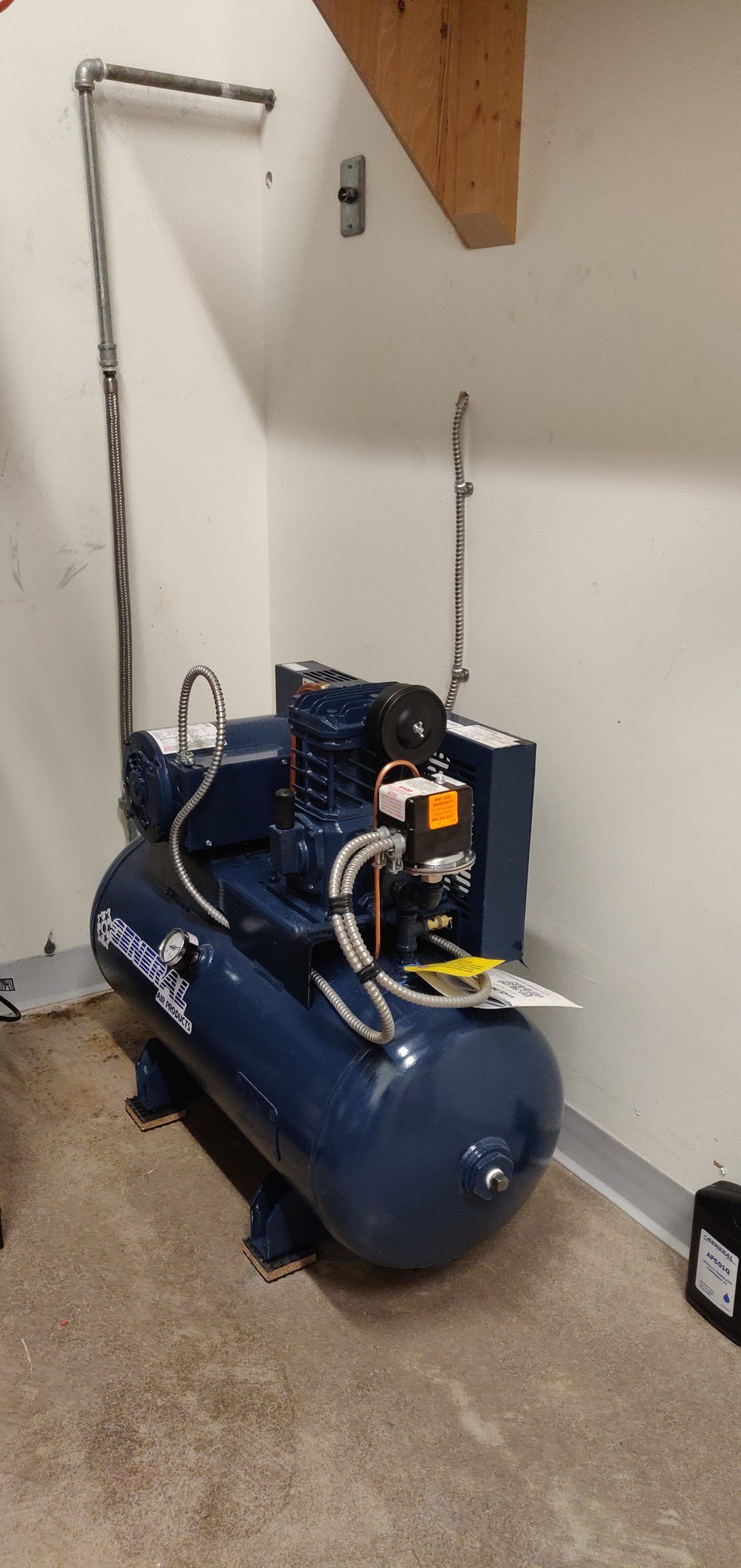 Blue air compressor in a workshop setting, with metal piping and exposed wooden beams.