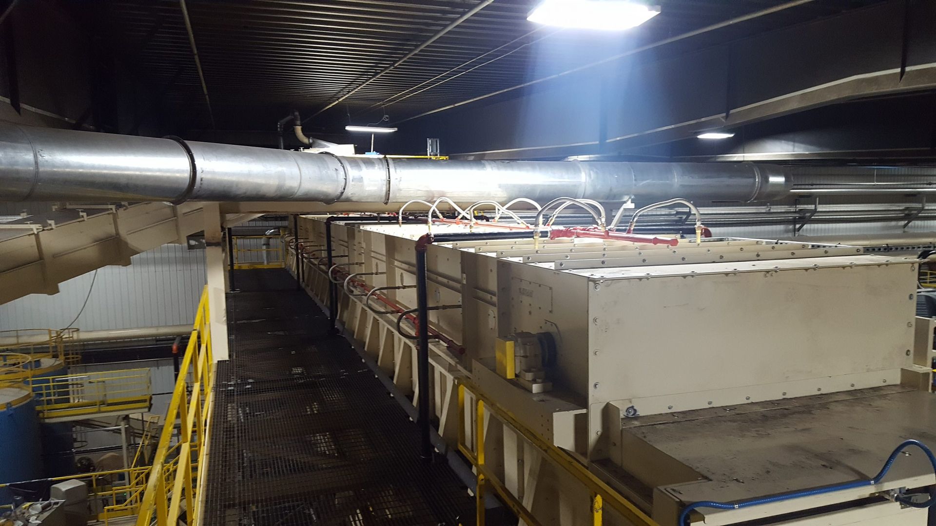 Factory interior with industrial machinery. Metal ductwork and overhead lights, long rectangular processing units with red pipes.