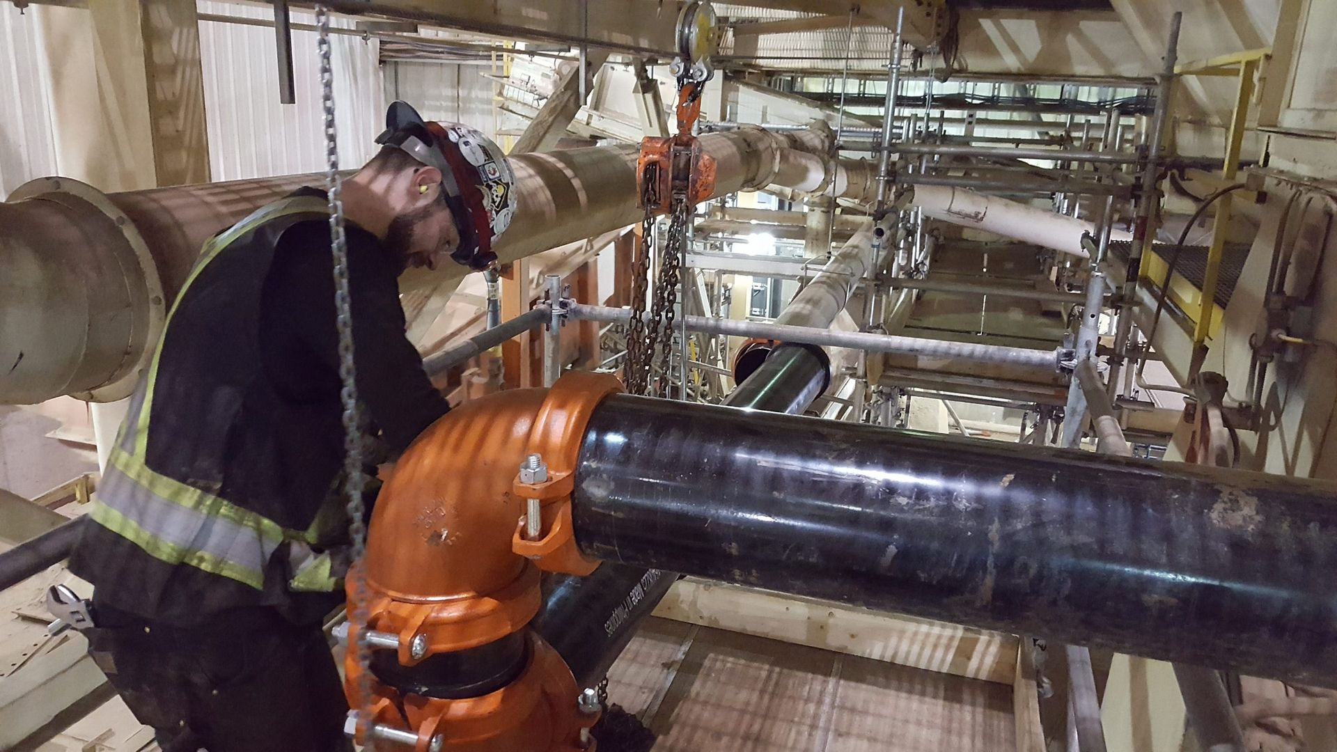 Worker in a hard hat inspecting industrial pipes on scaffolding, orange and black.