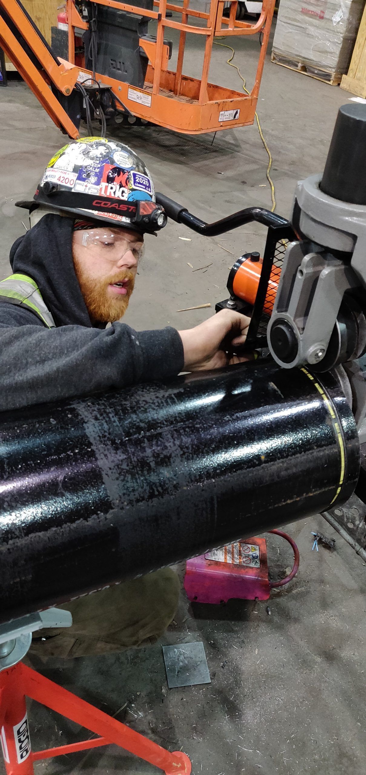 A person with a hard hat using a cutting tool on a large black pipe, indoors.
