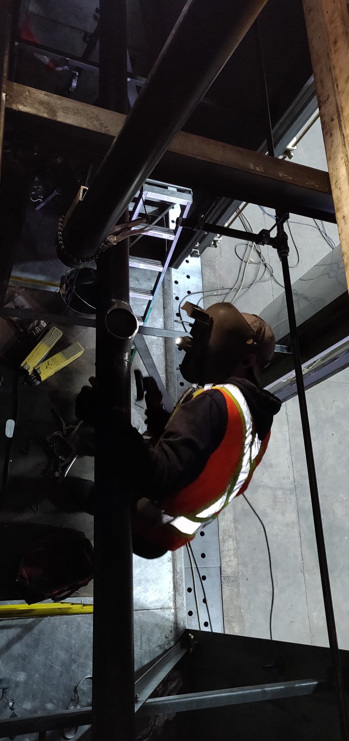 Worker in orange safety vest working on pipes, inside an industrial setting.