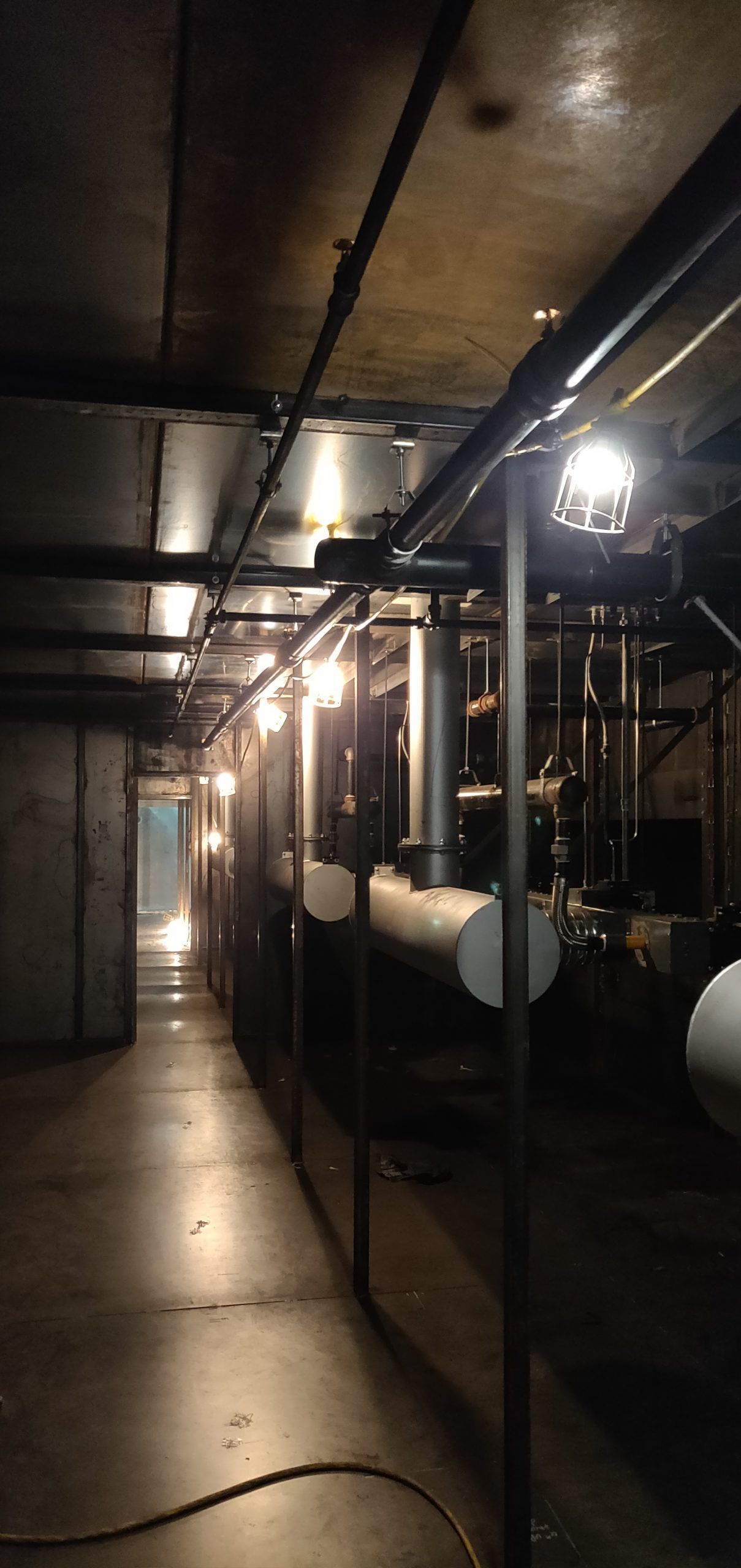 A dimly lit industrial space. Pipes and equipment line the walls. A long hallway leads to a bright opening.