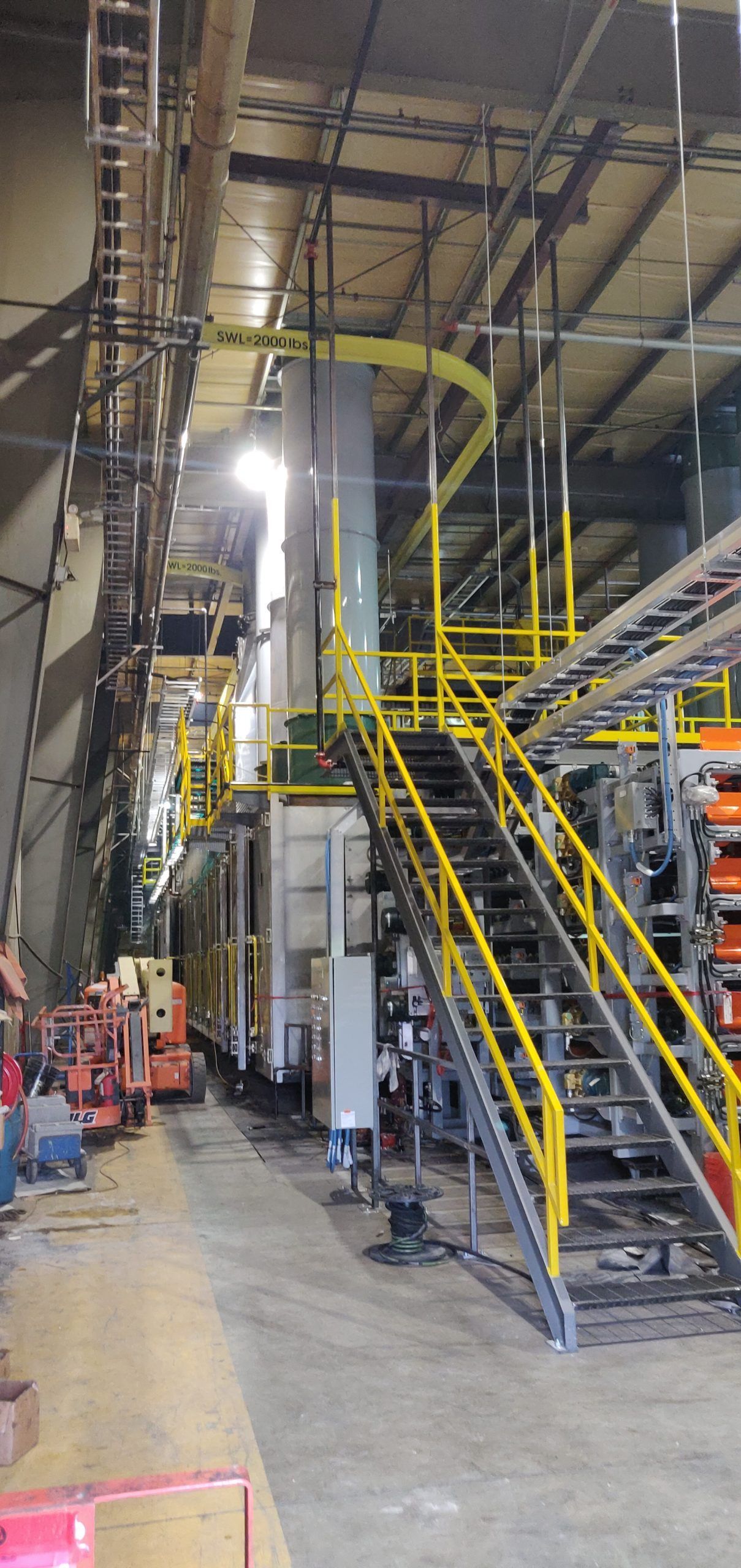 Industrial interior with machinery, stairs, and catwalks. Yellow railings, grey walls.