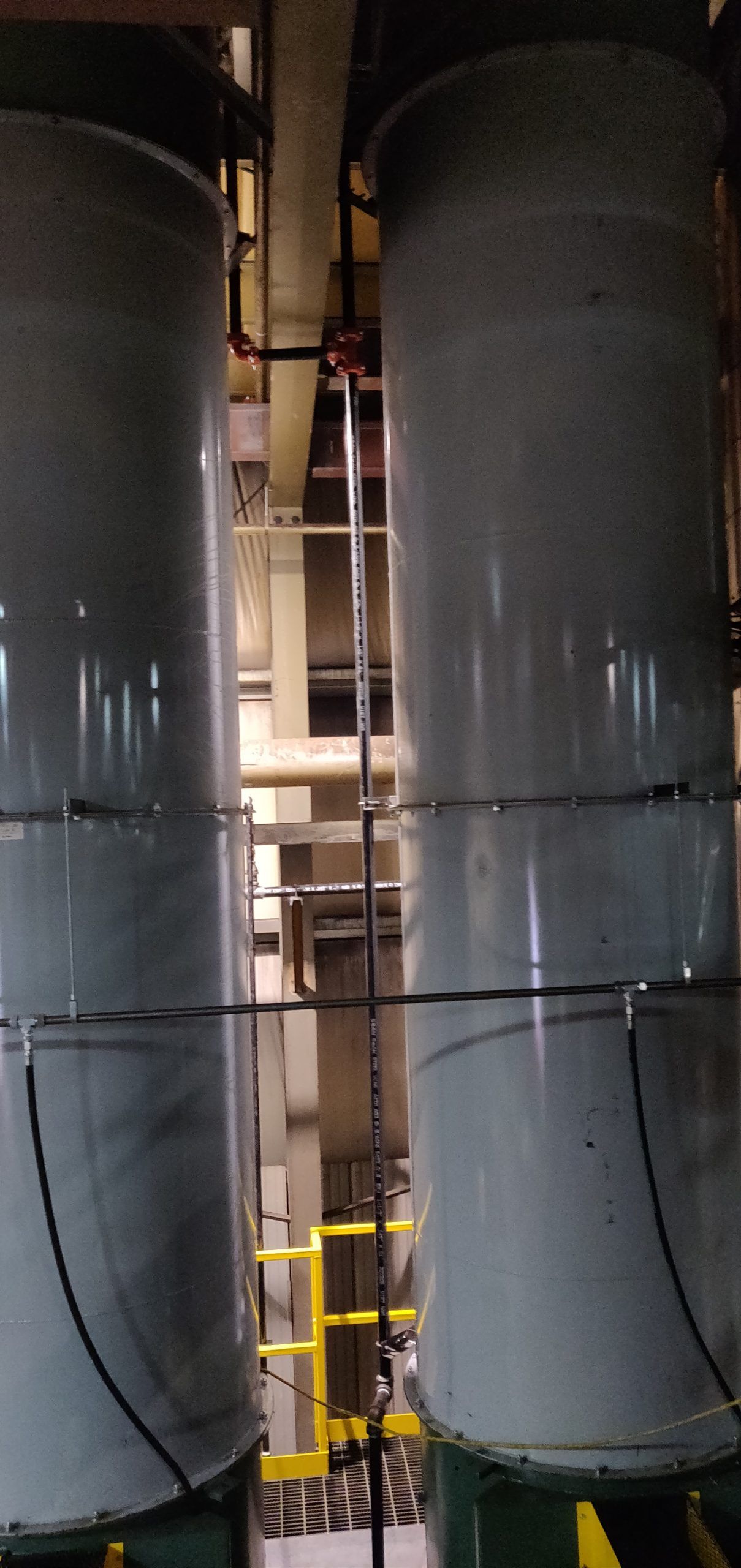 Two large cylindrical industrial tanks with piping and a yellow railing in the background.