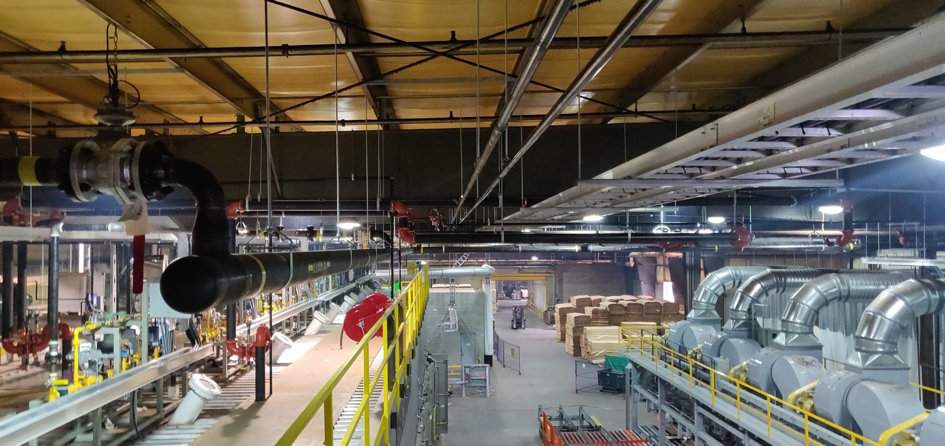 Industrial interior with pipes, machinery, and a metal roof. A yellow safety railing is in the foreground.