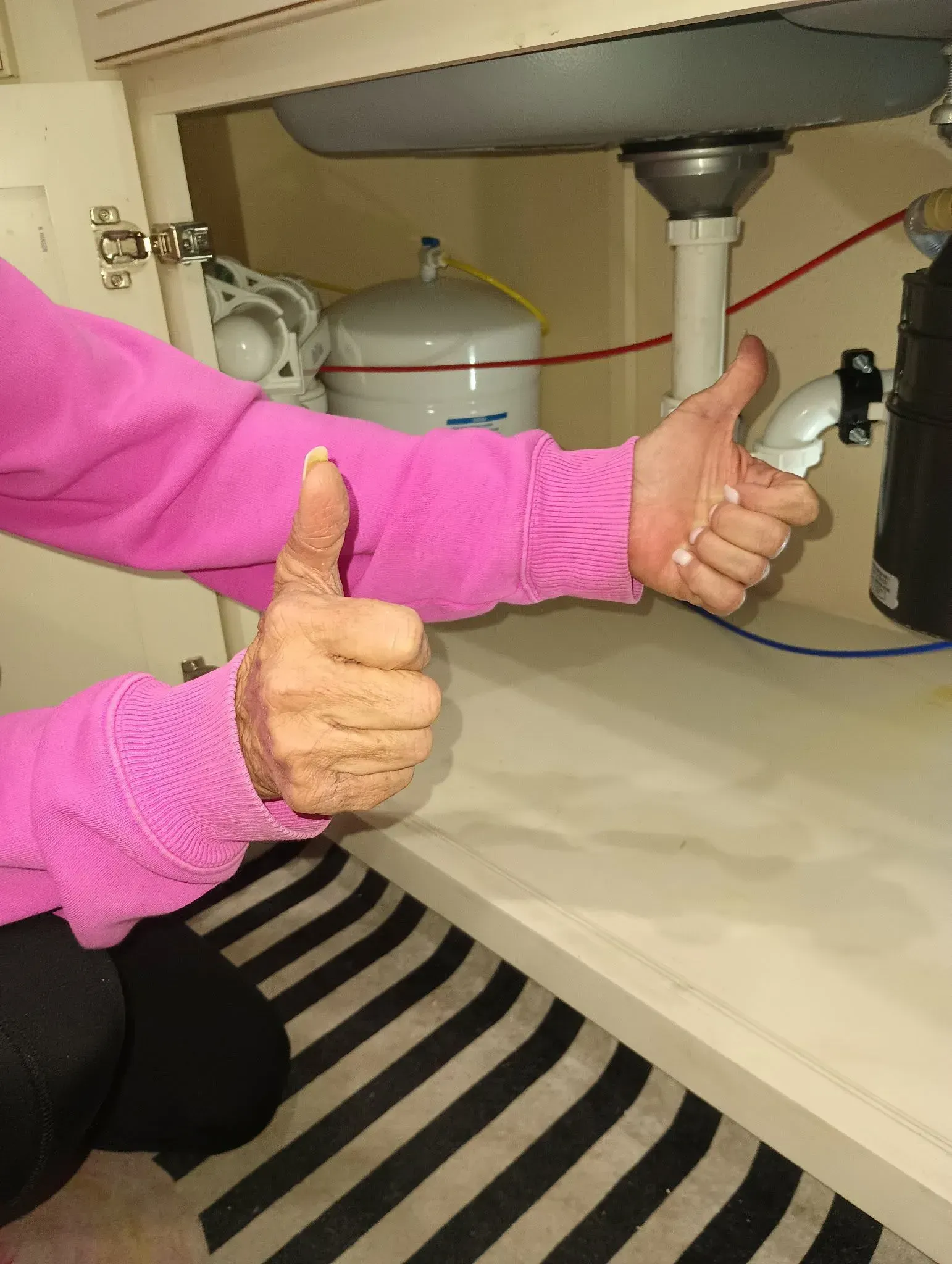 Person in pink sweater giving thumbs up under a sink cabinet, with plumbing visible.
