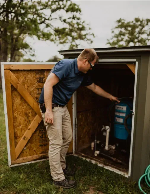 Man inspecting plumbing inside an open outdoor utility shed with a blue tank.