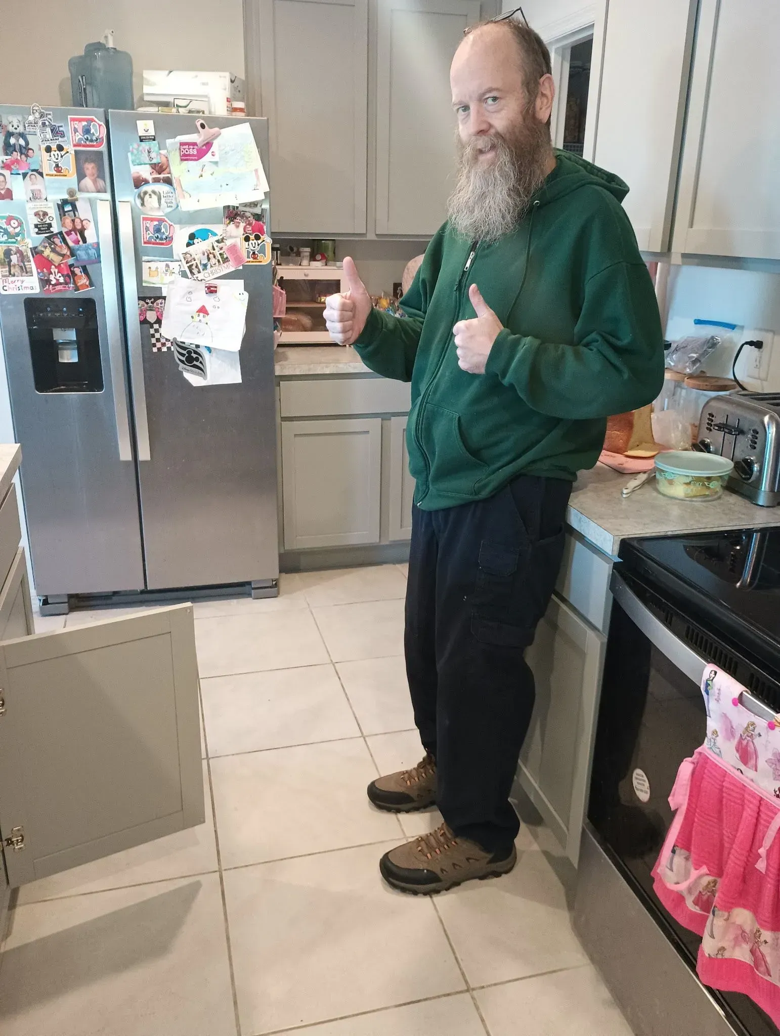 Man in green hoodie standing in a kitchen beside a fridge covered with magnets and notes.