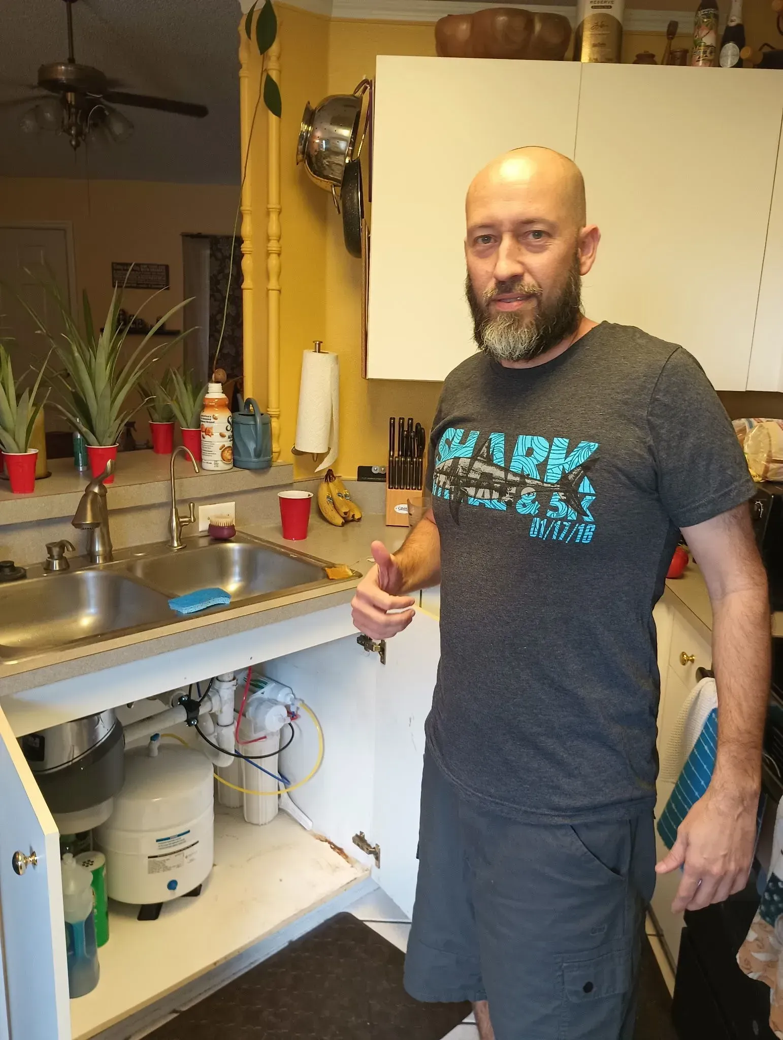 Man standing in a kitchen beside a sink, giving a thumbs-up, with cleaning items on the counter.