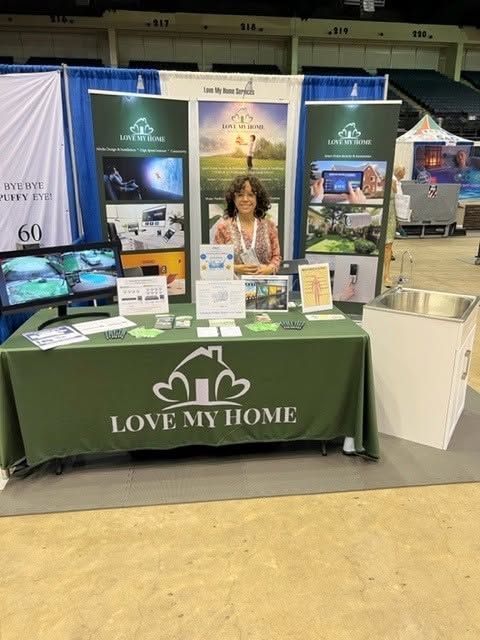 Trade show booth for “Love My Home” with green tablecloth, display boards, and a woman behind the table.
