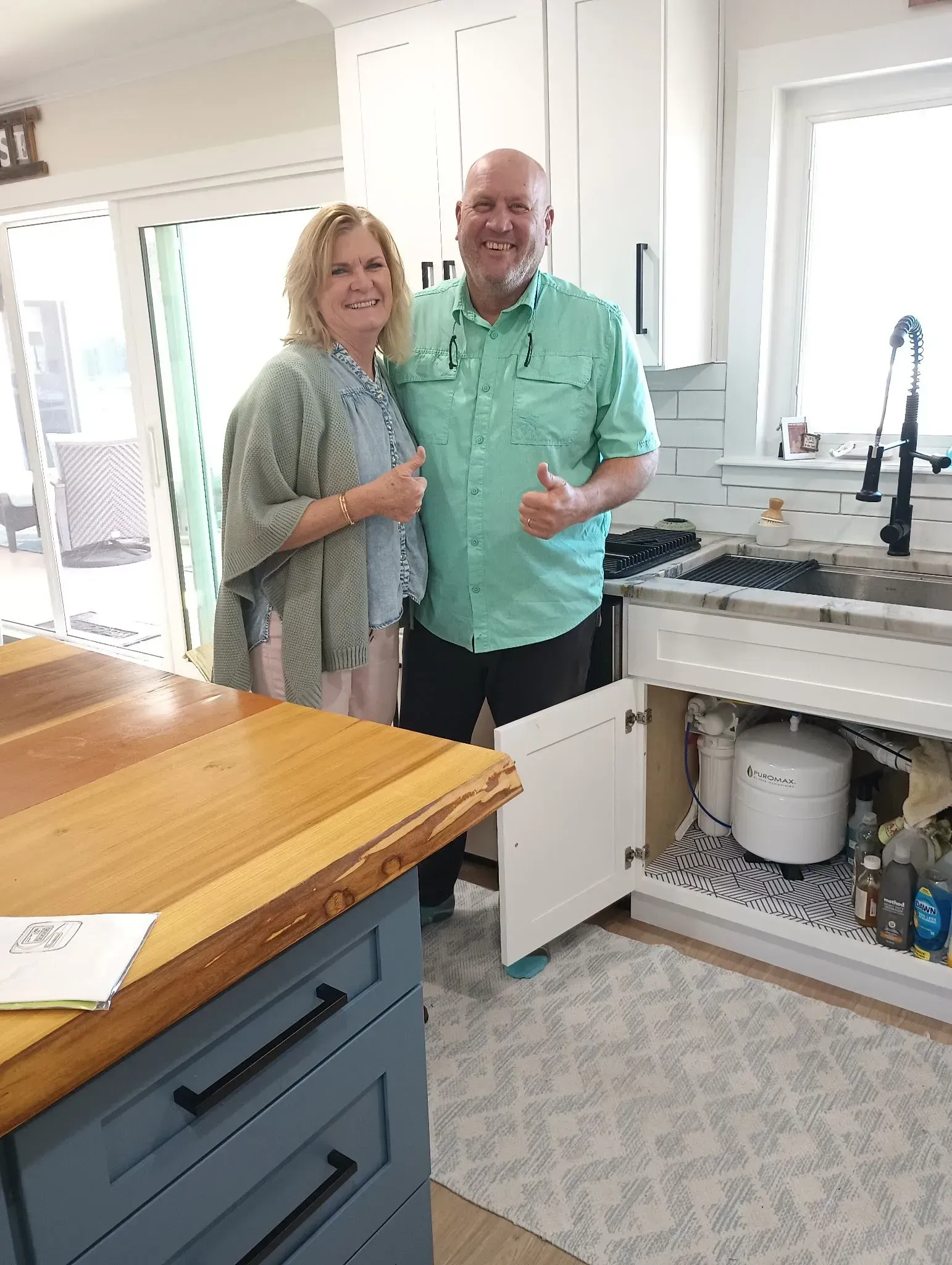 Smiling couple in a bright kitchen, standing by a sink and holding drinks.