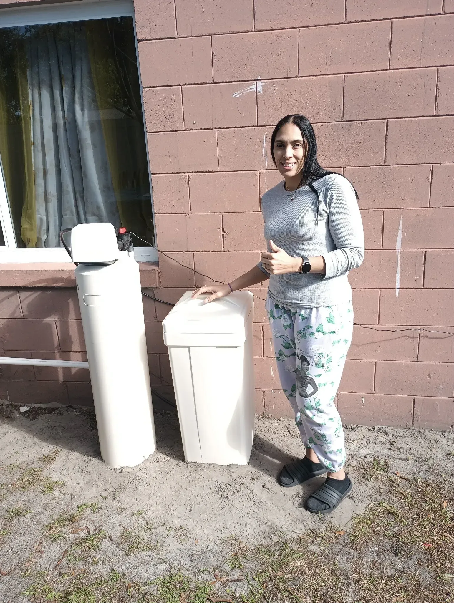 Person standing beside two white trash bins against a brick wall, near a window and curtain.