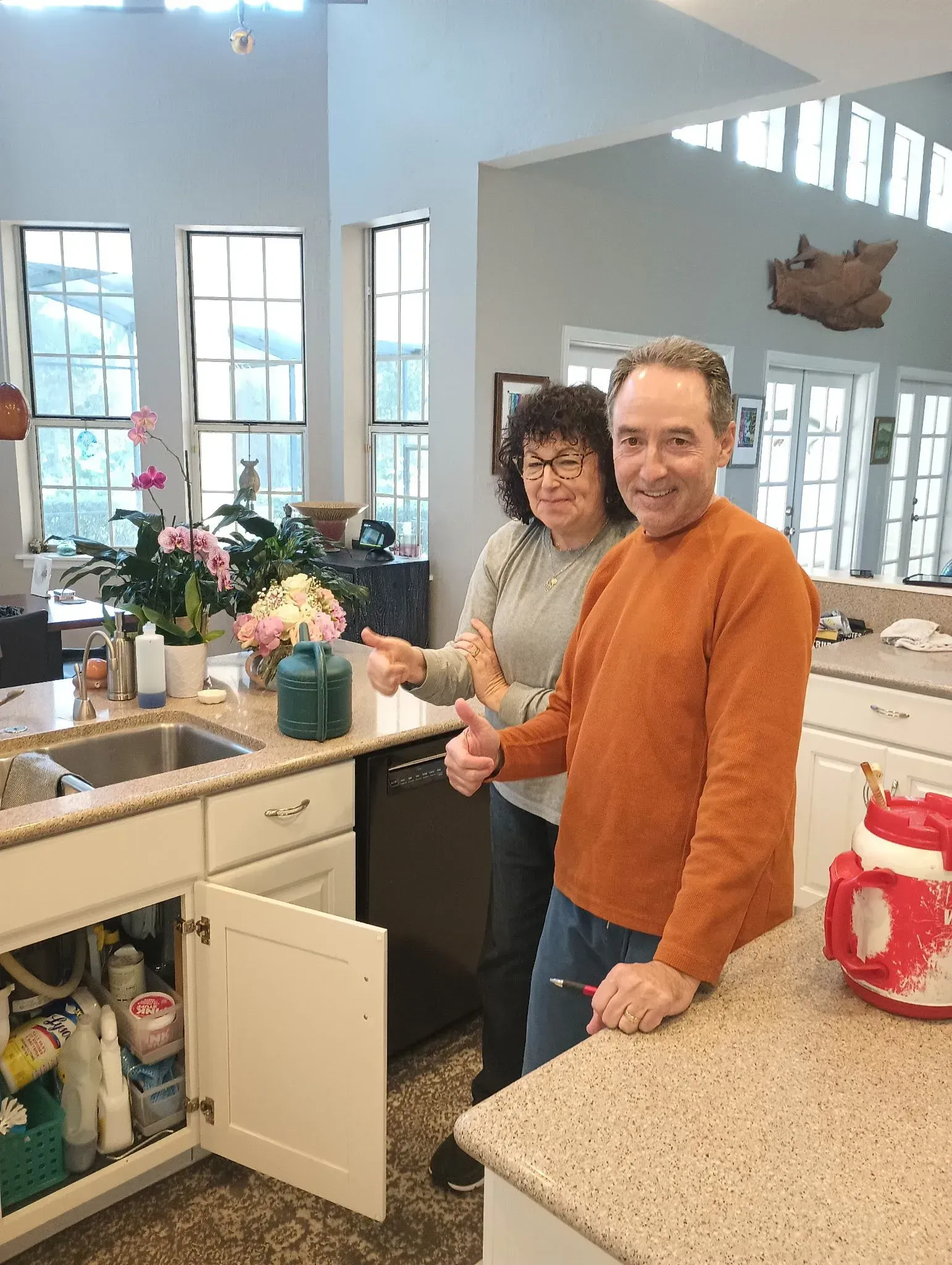 Two people smiling in a bright kitchen, standing by an open cabinet and countertop with flowers and a red kettle.