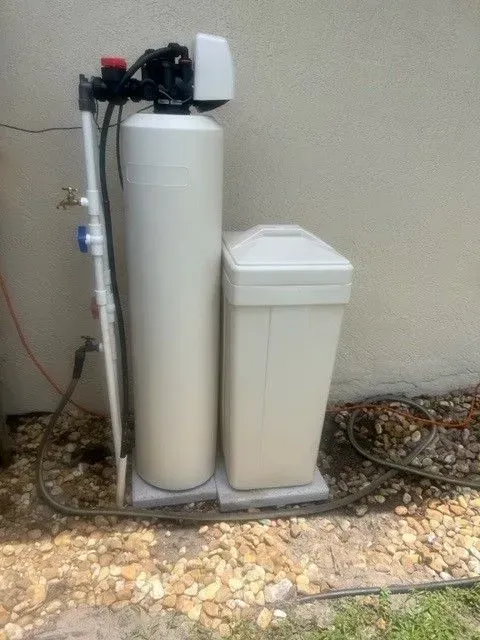 Water softener system with two white tanks and plumbing installed outdoors beside a wall.
