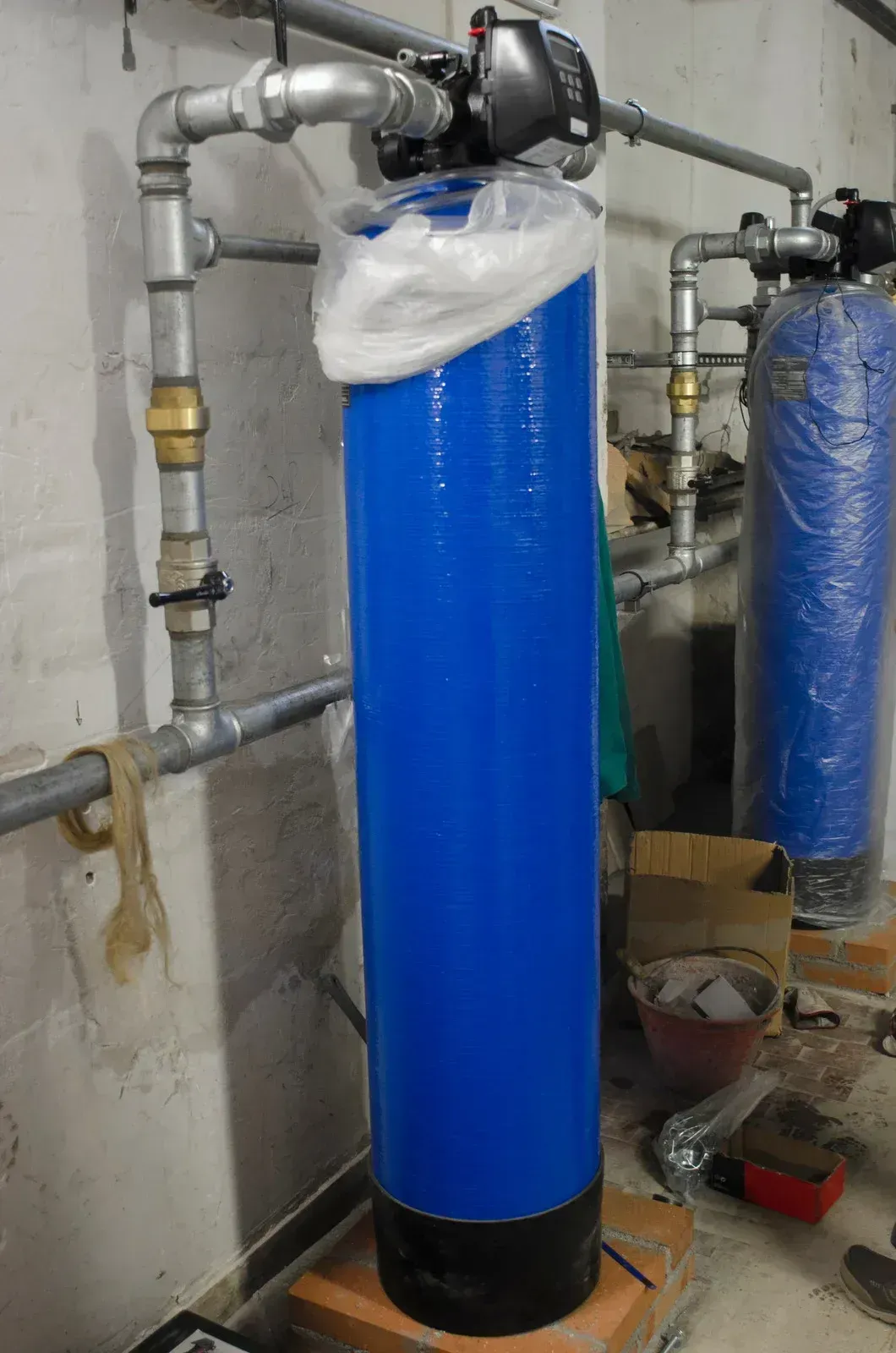Blue water filter tank with wrapped top, connected to gray pipes in a utility room