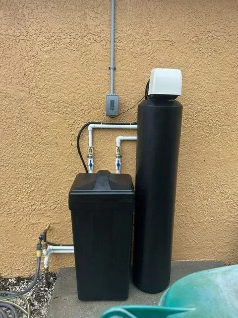 Two black water treatment tanks beside a stucco wall with exposed pipes and an electrical outlet