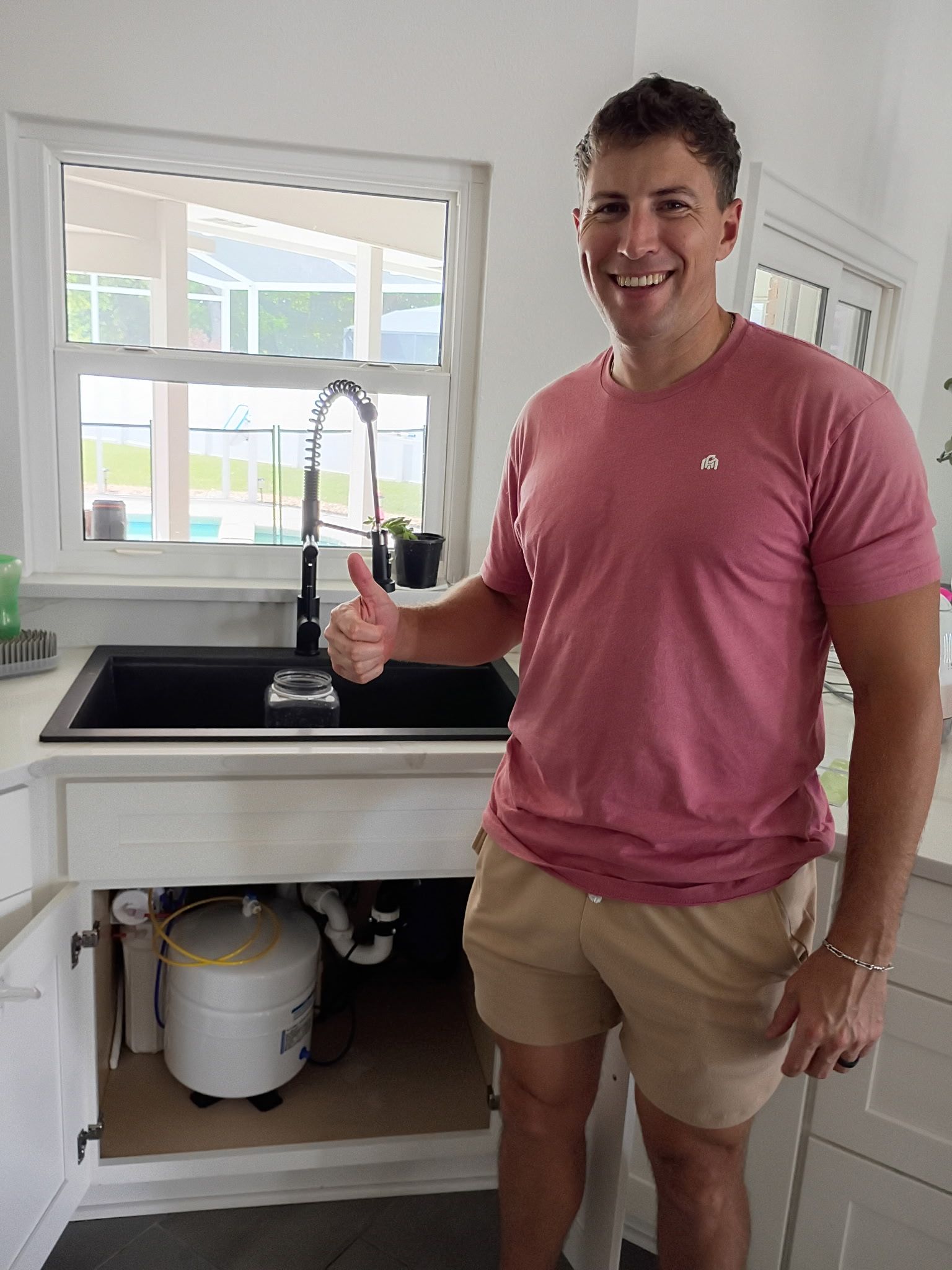 Man standing in a bright kitchen beside a sink, smiling and holding a bottle.