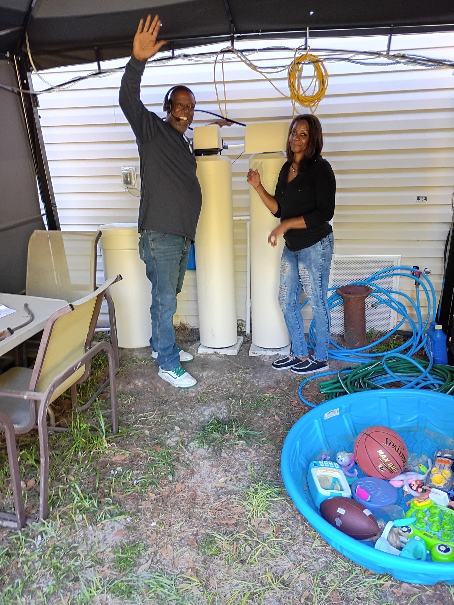 Two people standing in a garage beside large white tanks and a blue kiddie pool filled with toys.