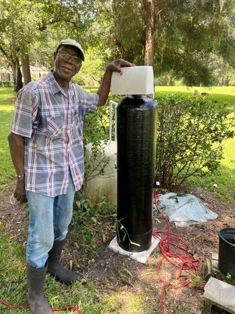 Man standing beside a tall black cylindrical tank outdoors, holding the top in a grassy yard.