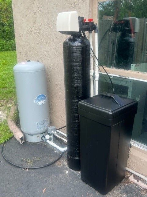 Outdoor water softener and filtration tanks beside a house window on a paved patio.