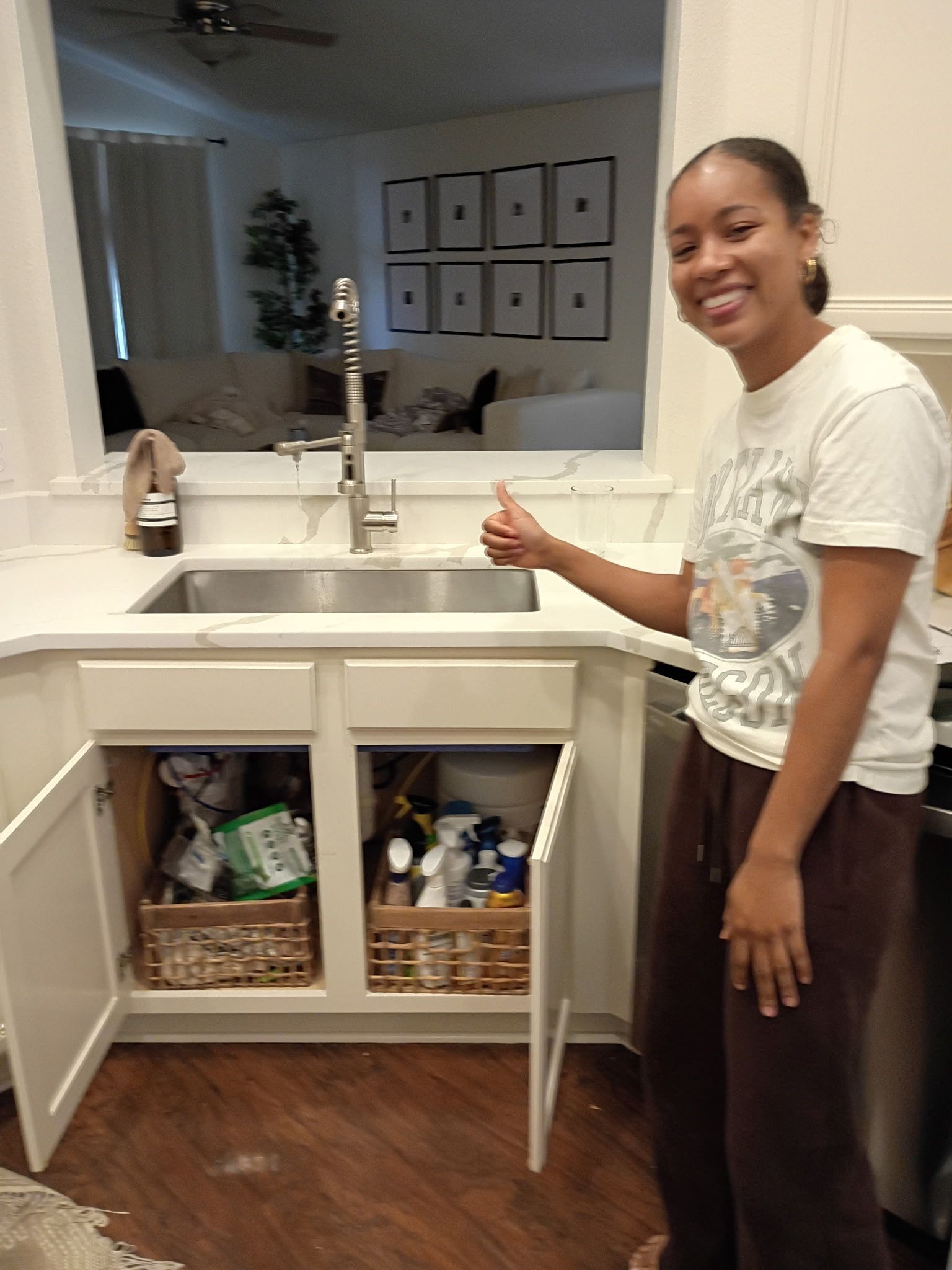 Woman standing by a white sink and open cabinet, smiling in a bright kitchen or bathroom.