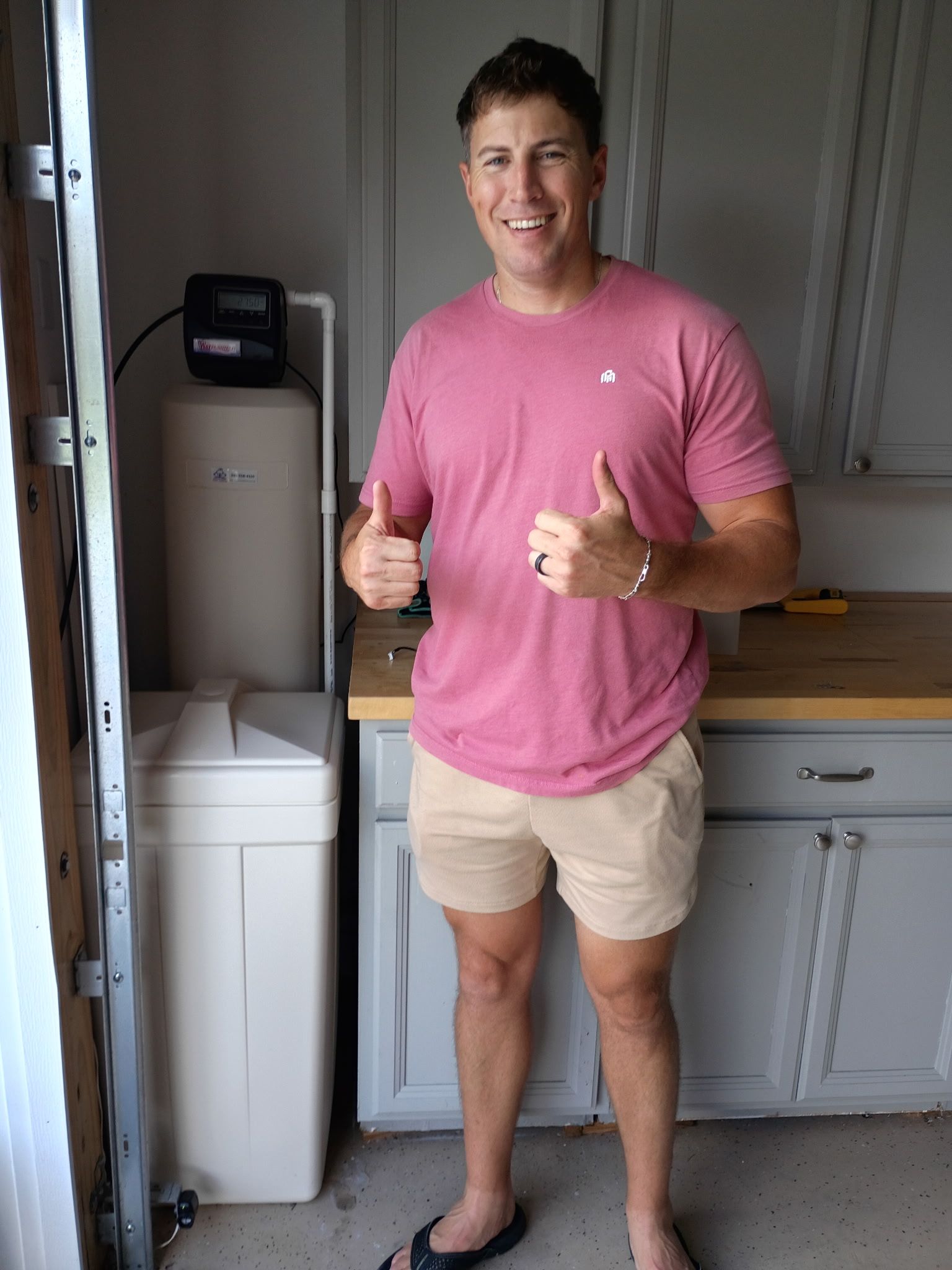 Smiling man in pink shirt and tan shorts giving thumbs up in a kitchen laundry area