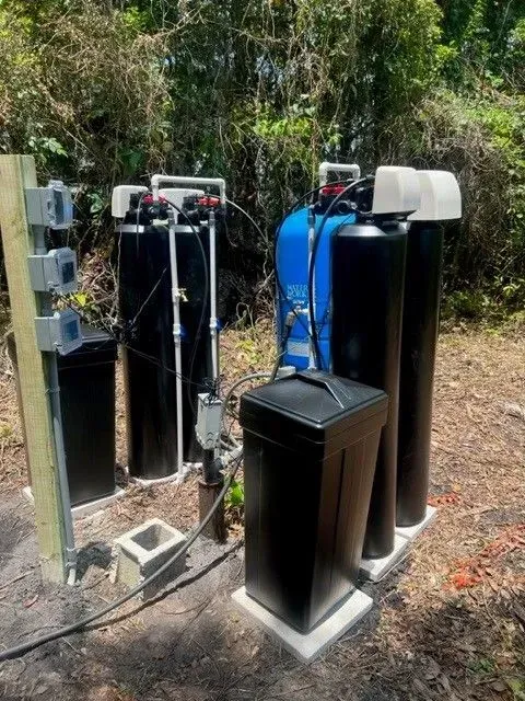 Outdoor water filtration system with black and blue tanks on a concrete pad beside trees