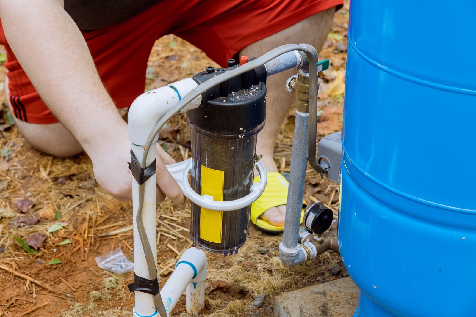 Person working on a black-and-yellow water filter connected to white pipes beside a blue barrel outdoors