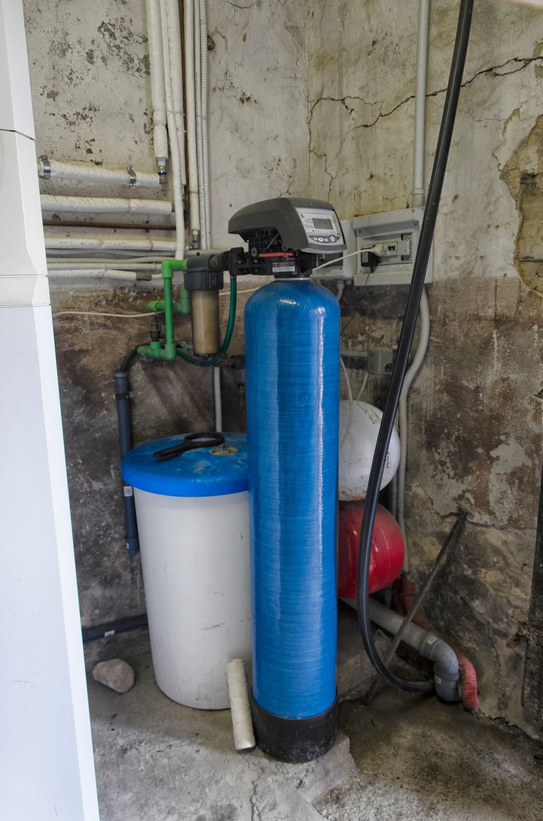 Blue water softener and white tank beside pipes and red pressure tanks in a basement utility area