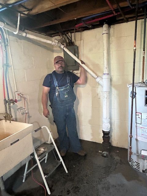 Worker in overalls standing in a basement beside exposed plumbing and a white utility pipe.