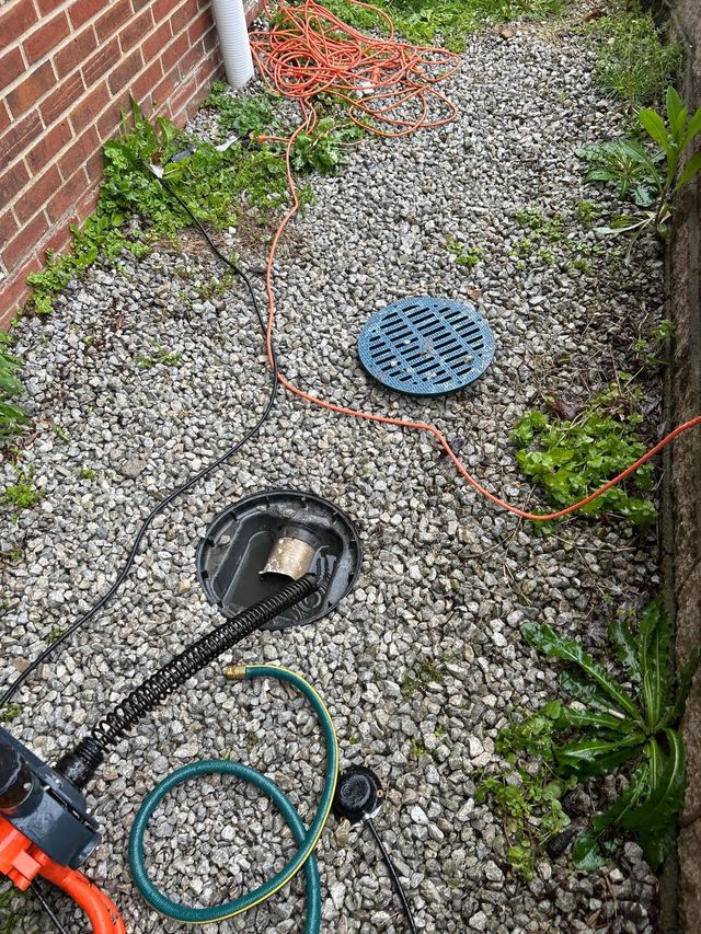 Outdoor gravel side yard with a blue drain cover and a cable reel near a brick wall and hose.