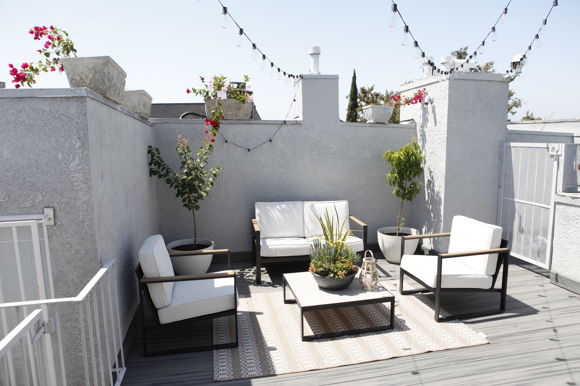 Rooftop patio with white furniture, string lights, and potted plants on a sunny day.