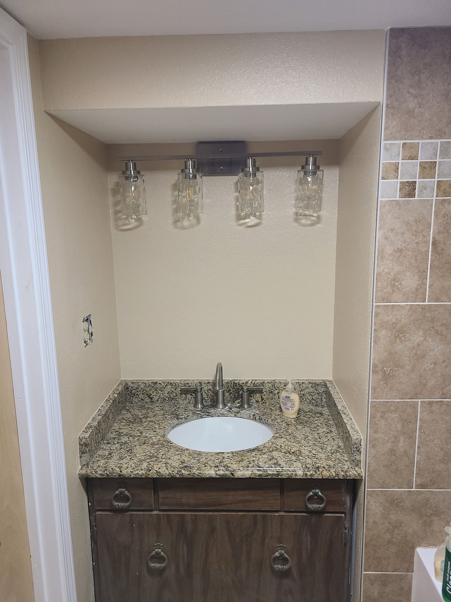 A bathroom vanity with a speckled granite countertop, white sink, wood cabinet, and a four-light mason jar fixture.
