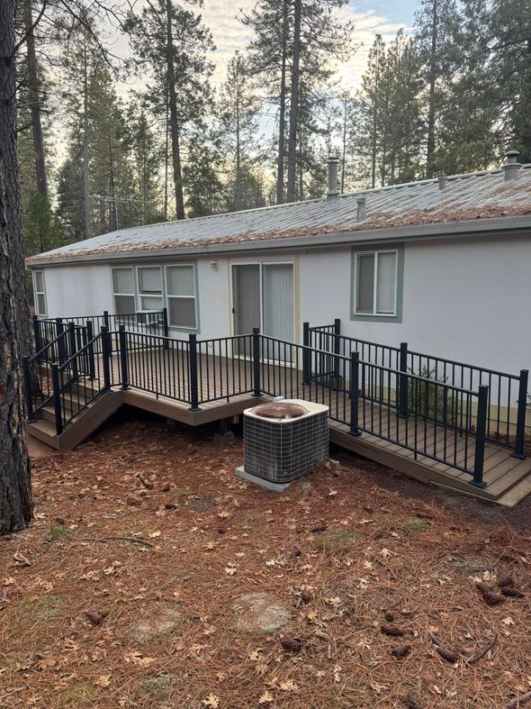 A single-story home surrounded by pine trees, featuring a wooden deck with black railings and an attached handicap ramp.